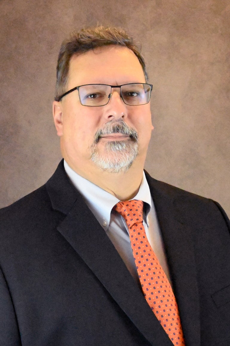 A man with glasses wears a black suit jacket, light blue button up, and orange patterned tie.