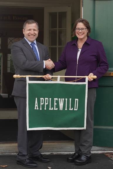 A man in a dark grey suit, light blue button down, and dark blue tie shakes hands with a woman in a purple jacket and dark grey pants while they both hold a green sign that reads "Applewild"