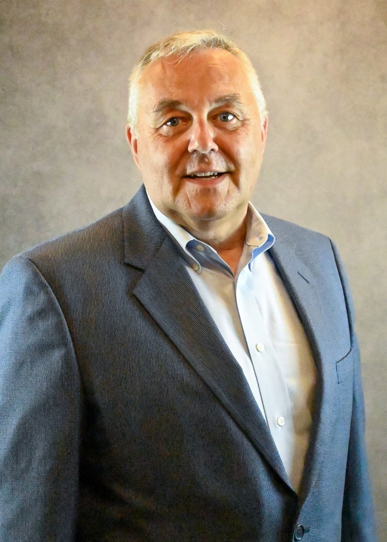 A headshot of a man with grey hair weraing a dark grey suit jacket and light blue button up