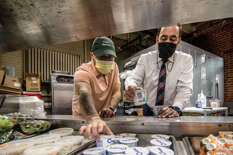 A man in a green baseball cap and a man in a white button down wear masks reach for yogurts in containers in a top open kitchen refrigerator.