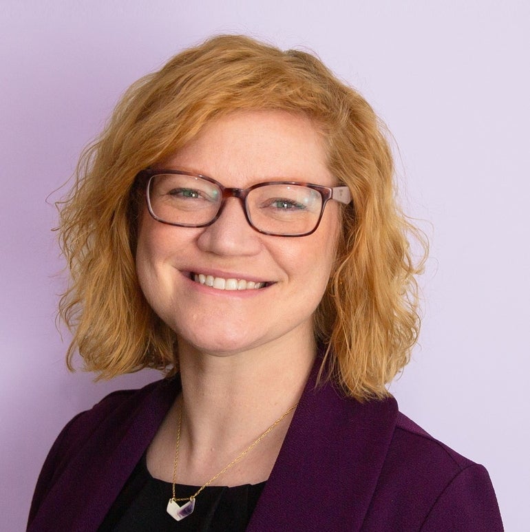 A headshot of a woman with wavy red hair wearing a glasses, a dark purple suit jacket, a black top, and a white and purple pendant necklace