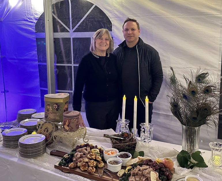 A man and woman, both wearing black, stand together behind a long white table with food, plates, and candles on it inside a white tent.