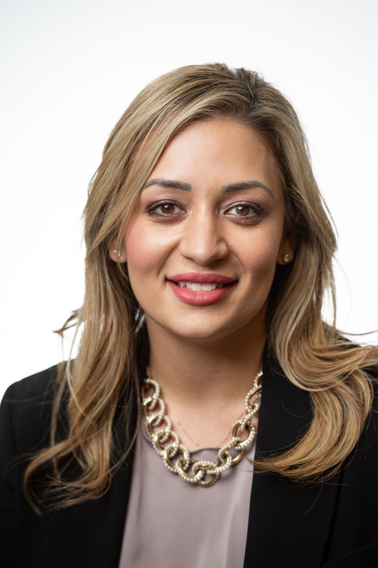 A woman with long blonde hair wears a black blazer, a grey/mauve top, and a gold chain necklace.