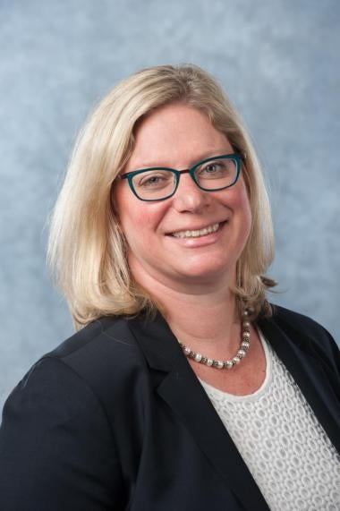 A woman with shoulder length blonde hair wears a pearl necklace, a black blazer, and a white eyelet top.