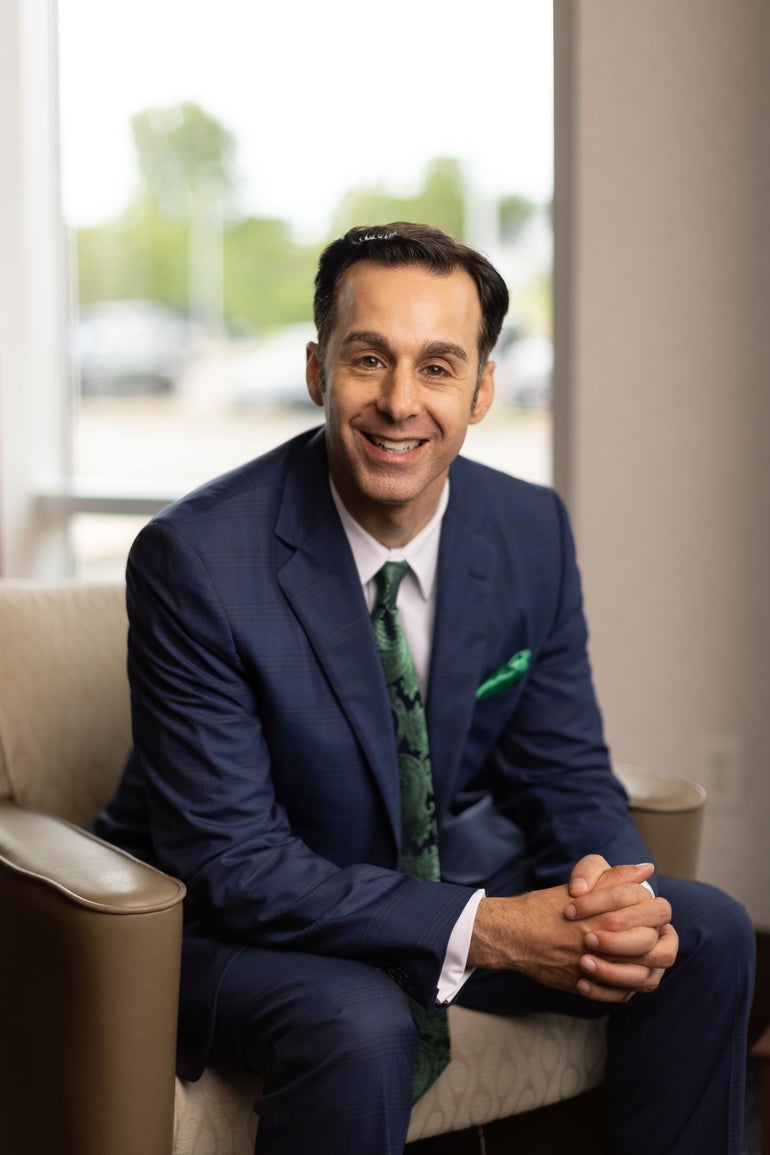 A man with short brown hair wears a dark blue suit, white button down, and green paisley tie while sitting on a beige chair and leaning forward with his elbows on his legs and hands folded together.