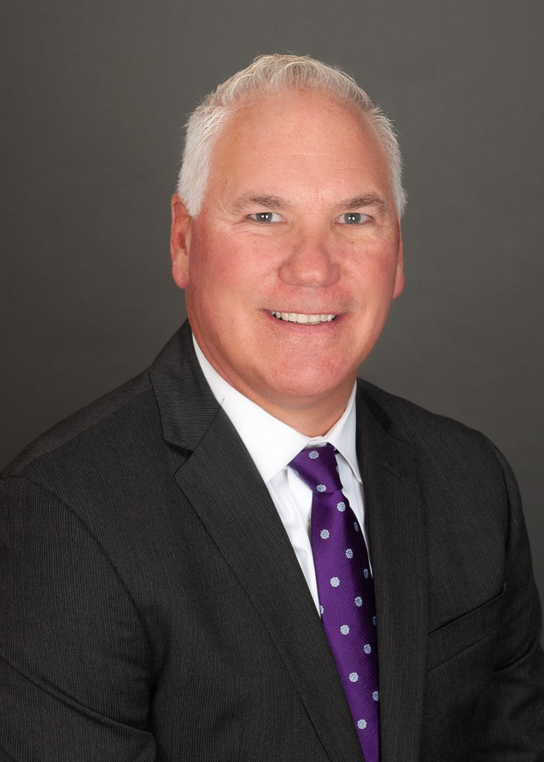 A man with grey hair wears a black suit jacket, a white button down, and a purple tie with white polka dots.