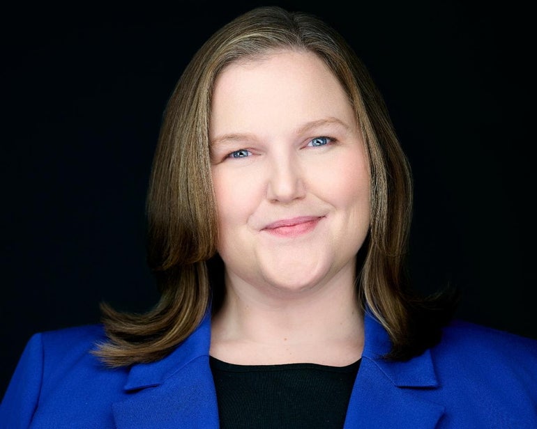 A woman with shoulder-length brown hair and blonde highlights wears a blue blazer and black top.