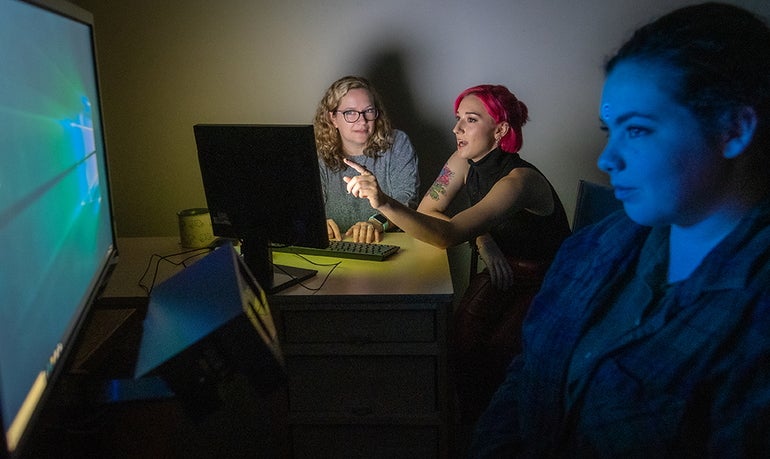 Three women, one with blonde hair, one with pink hair, and one with brown hair, sit in a dark room looking at computer screens.