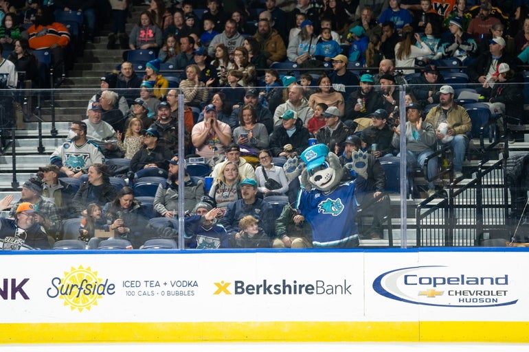 A dog mascot pushes itself up against the glass of a hockey rink, with a crowd behind it 