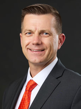 A man with short brown hair wears a black suit jacket, a white button down, and an orange-patterned tie.