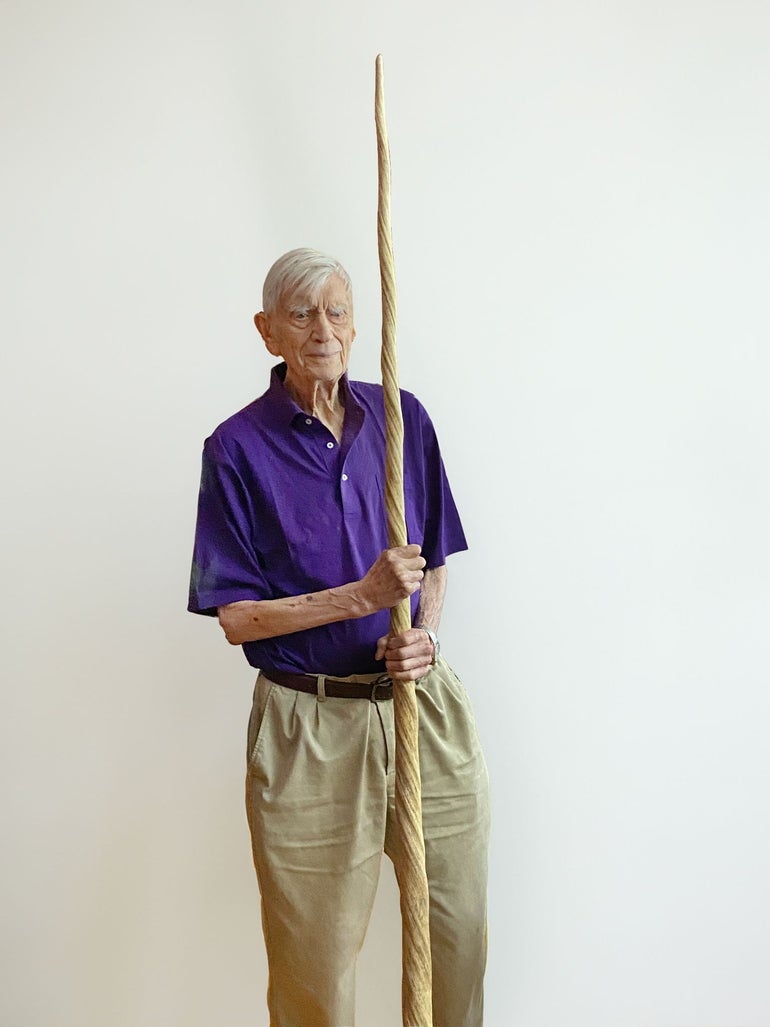 A man with white hair wears a purple polo and tan khakis while holding a long narwhal tusk.