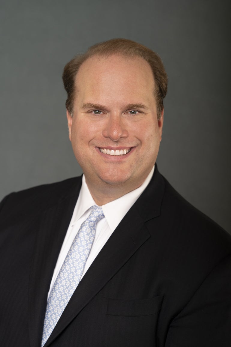 A man with light brown hair smiles for the camera wearing a black suit jacket, a light blue floral patterned tie, and a white button down.