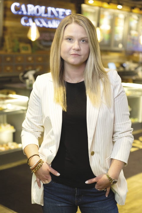 A woman with blond hair wearing a a black shirt and white jacket stands in front of a sign, which says "Caroline's Cannabis".