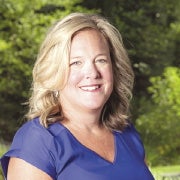 A woman with blonde hair wears a blue v-neck blouse in front of green trees.