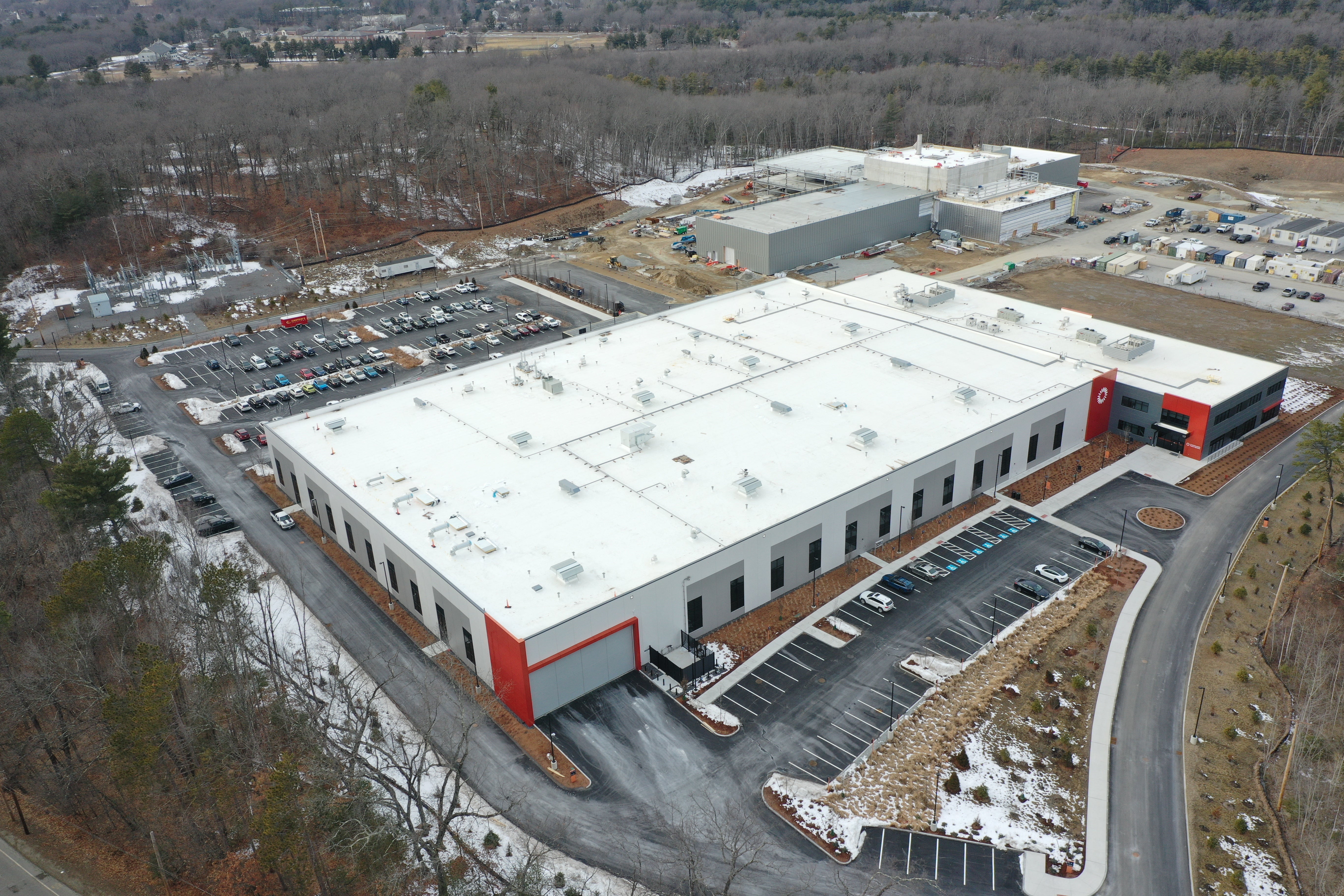 An aerial view of a large industrial building with a white roof and a parking lot behind