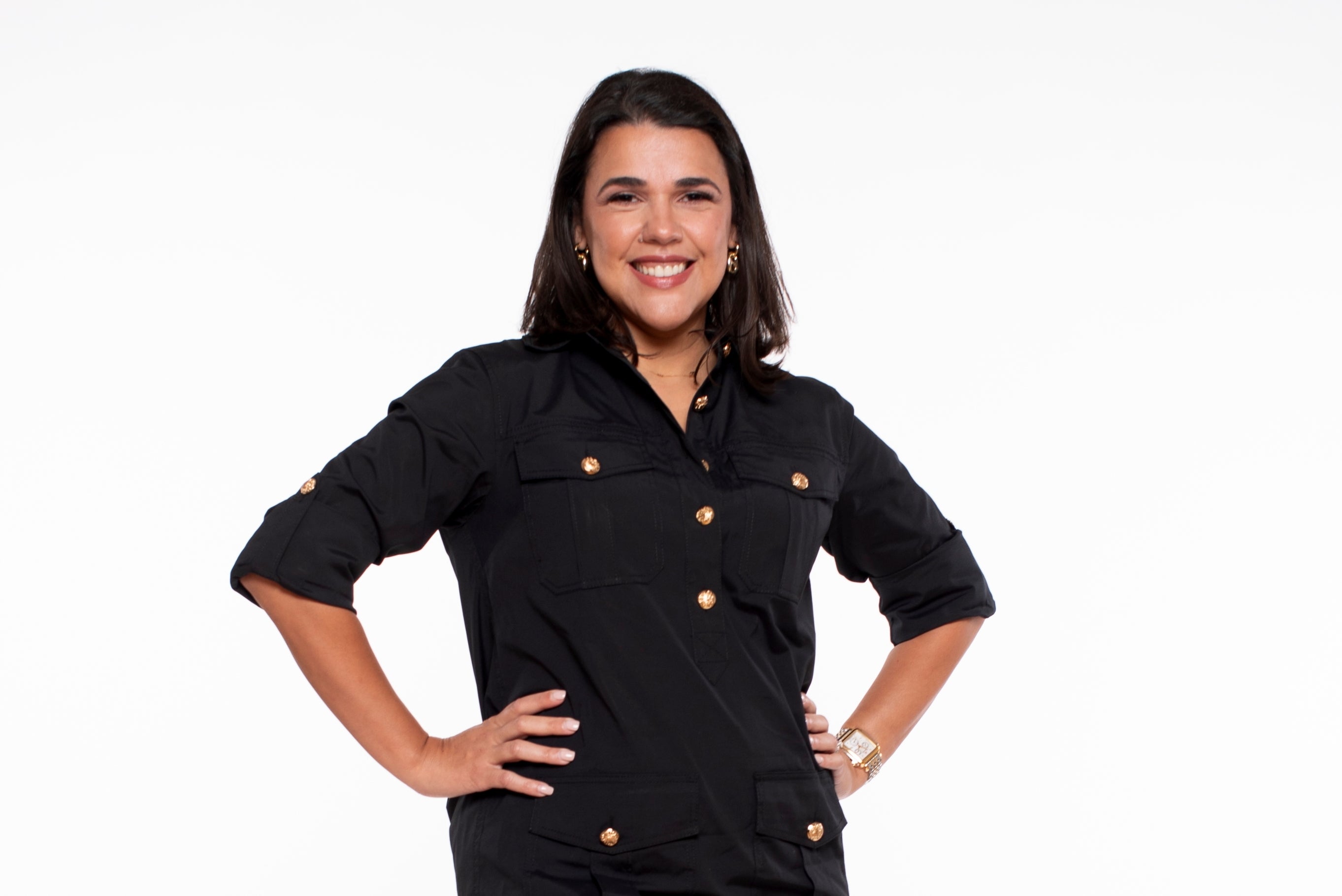 A woman with shoulder length dark hair wears a black dress with gold buttons with her hands on her hips.