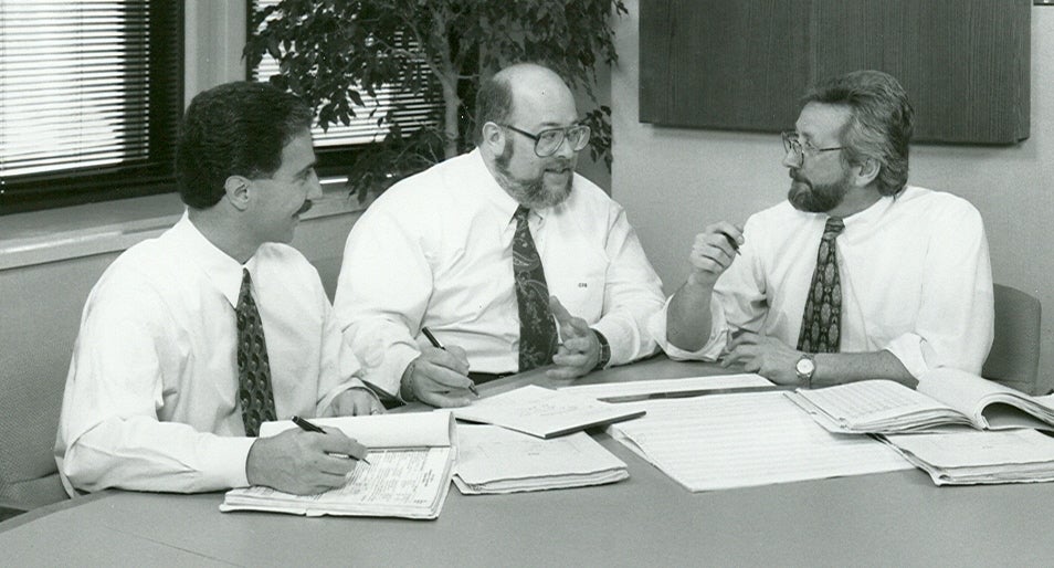 A black and white photo of three men in white button downs and patterned ties sitting at a table with papers and notepads on the table.