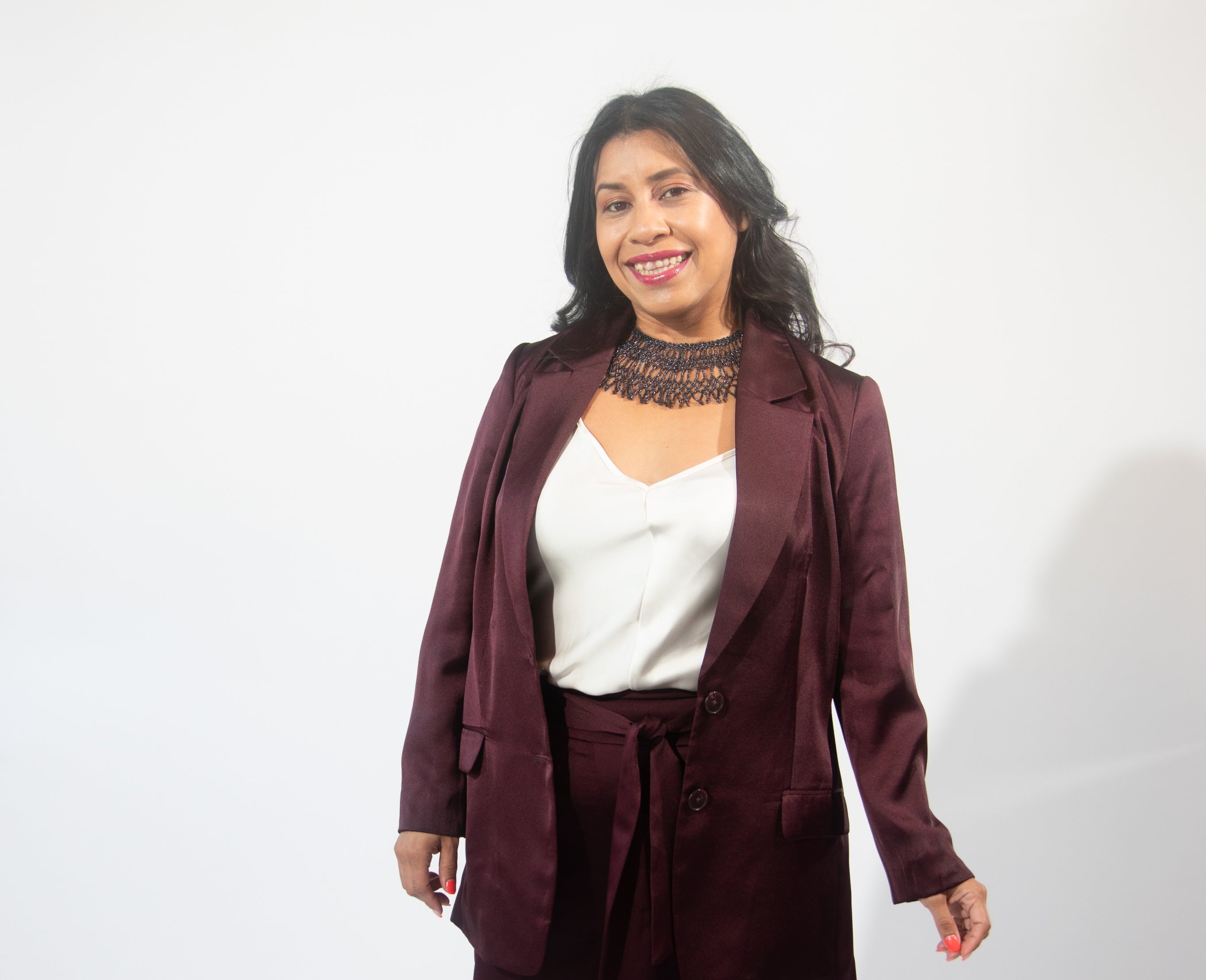 A woman with long black hair wears a black beaded necklace, a white satin top, and maroon satin suit jacket and pants.