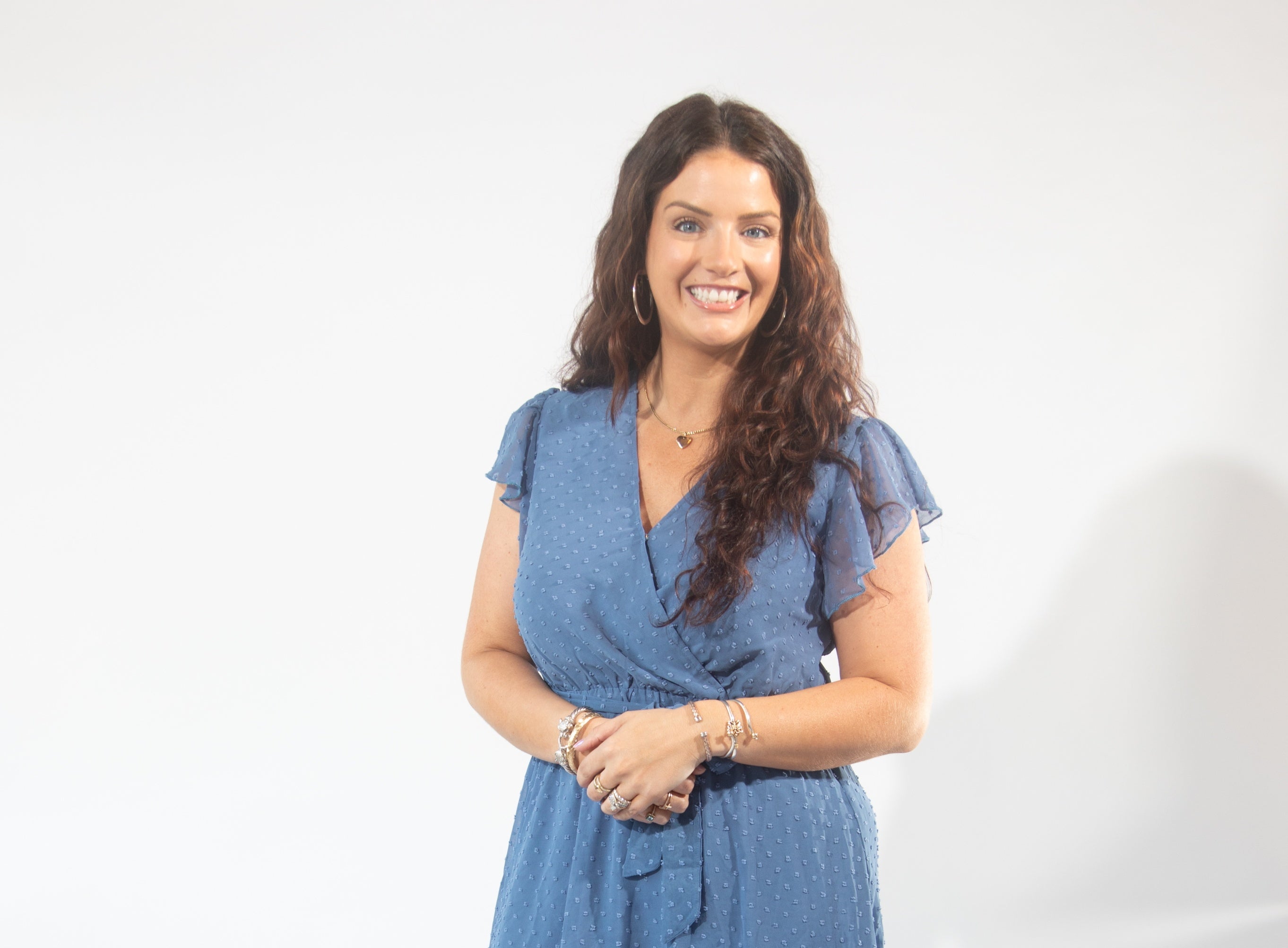 A woman with long curly brown hair wears a blue dress, hoop earrings, and a silver heart necklace.