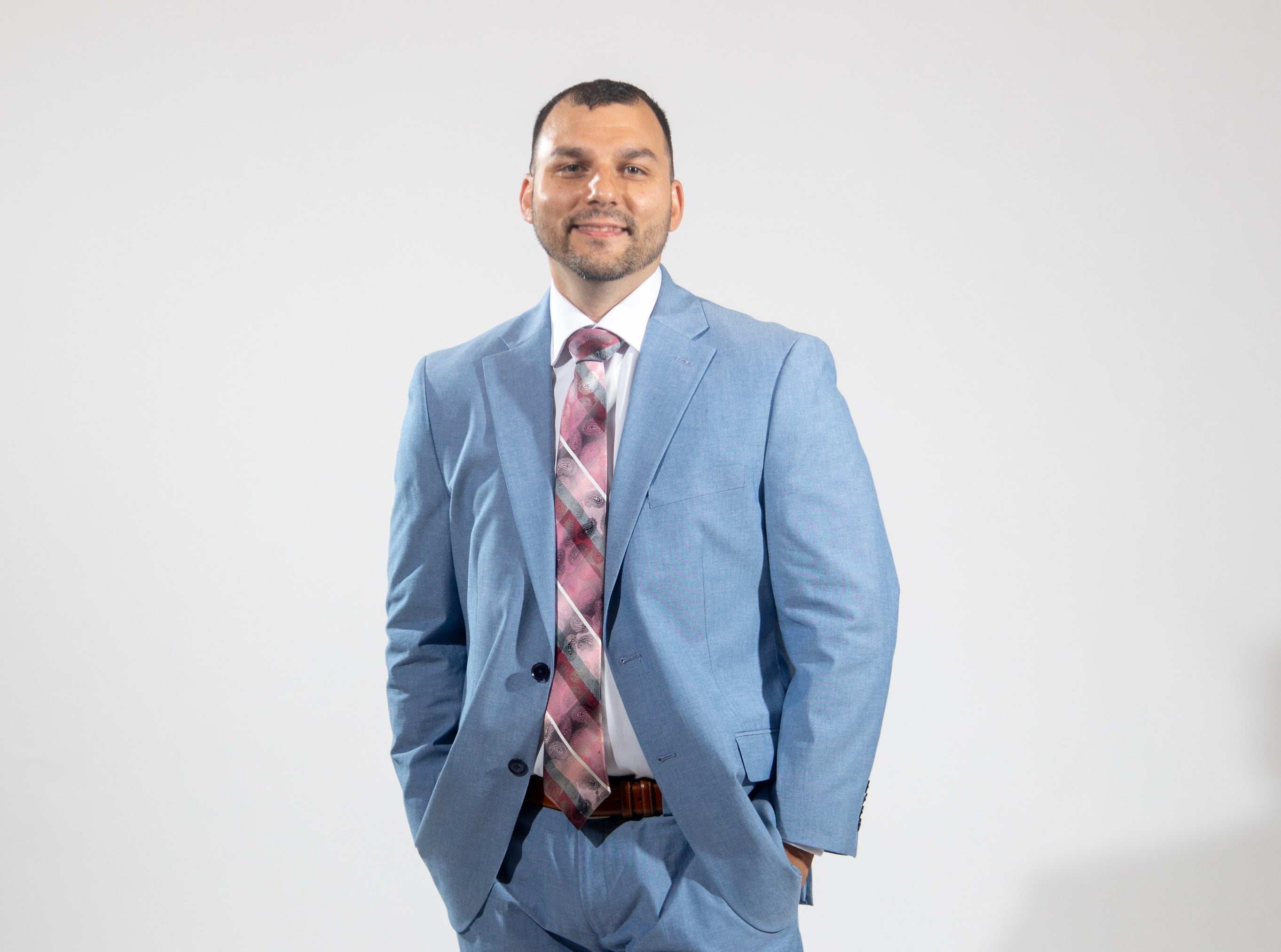 A man wears a light blue suit jacket and pants, a brown belt, a white button down, and a pink, grey, and white patterned tie with his hands in his pockets.