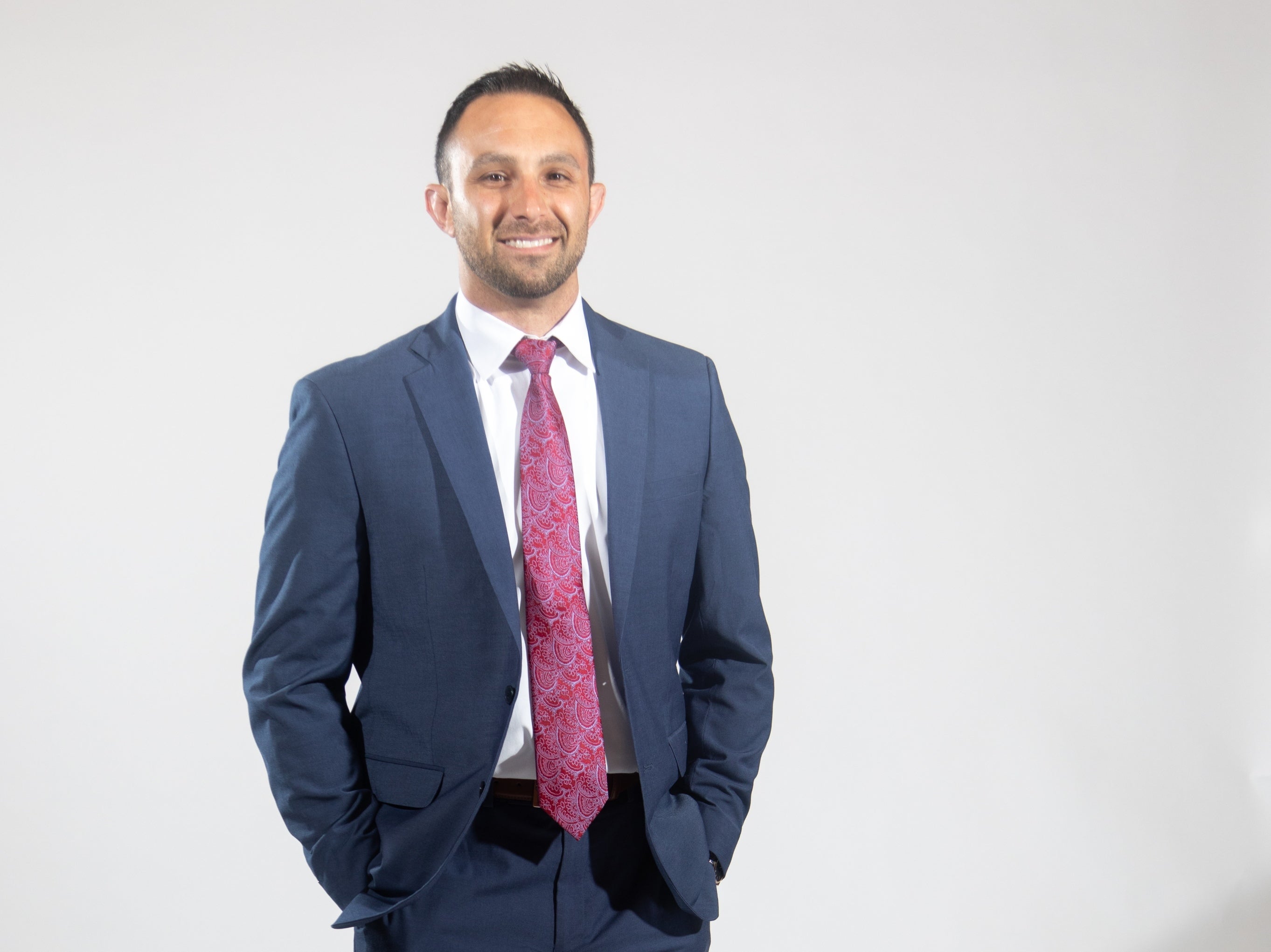 A man wears a blue suit jacket and pants, a white button down, and a purple and red paisley tie.