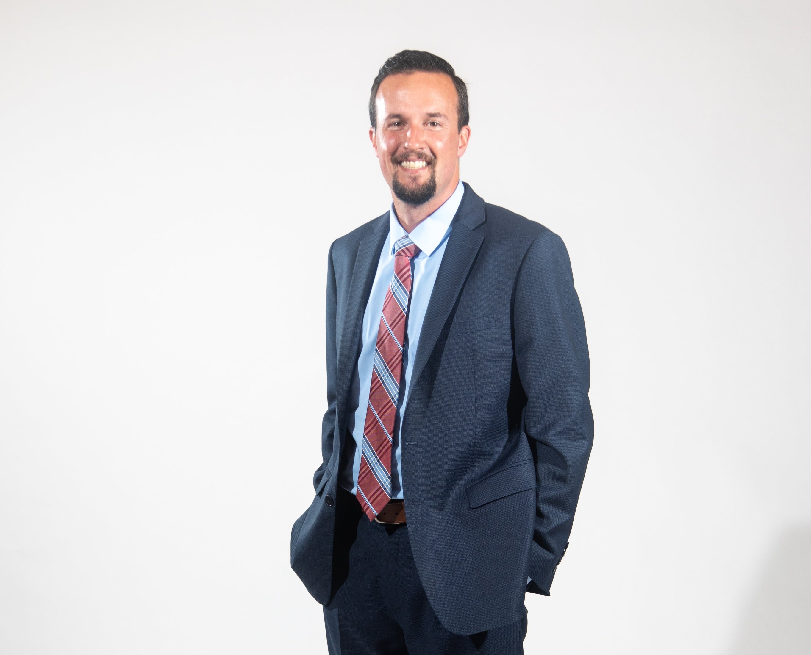 A man with short brown hair and a goatee wears a dark blue suit jacket and pants, a light blue button down, and a red and light blue plaid tie with his hands in his pockets.