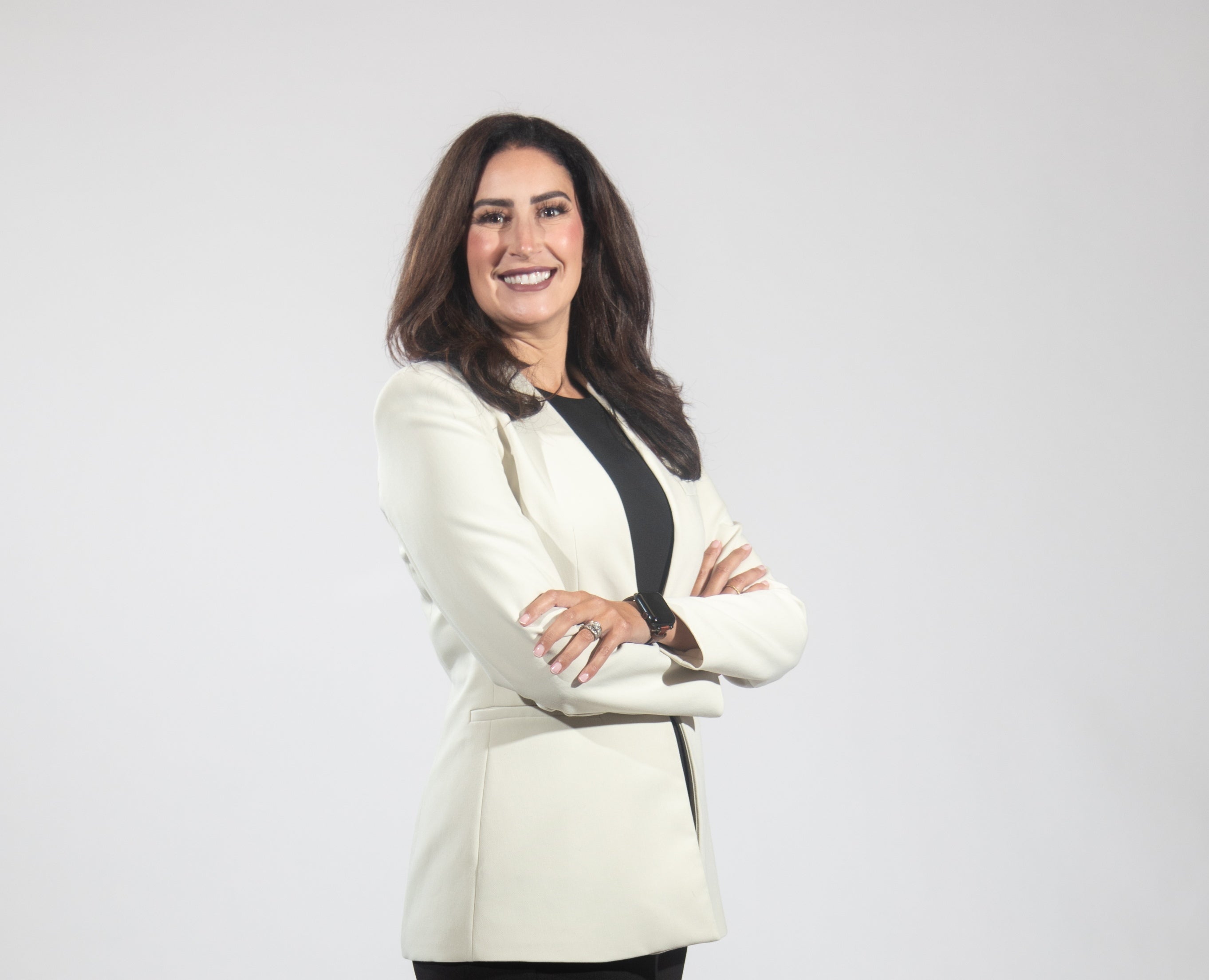 A woman with long brown hair wears a black top and white blazer, standing with her arms folded.