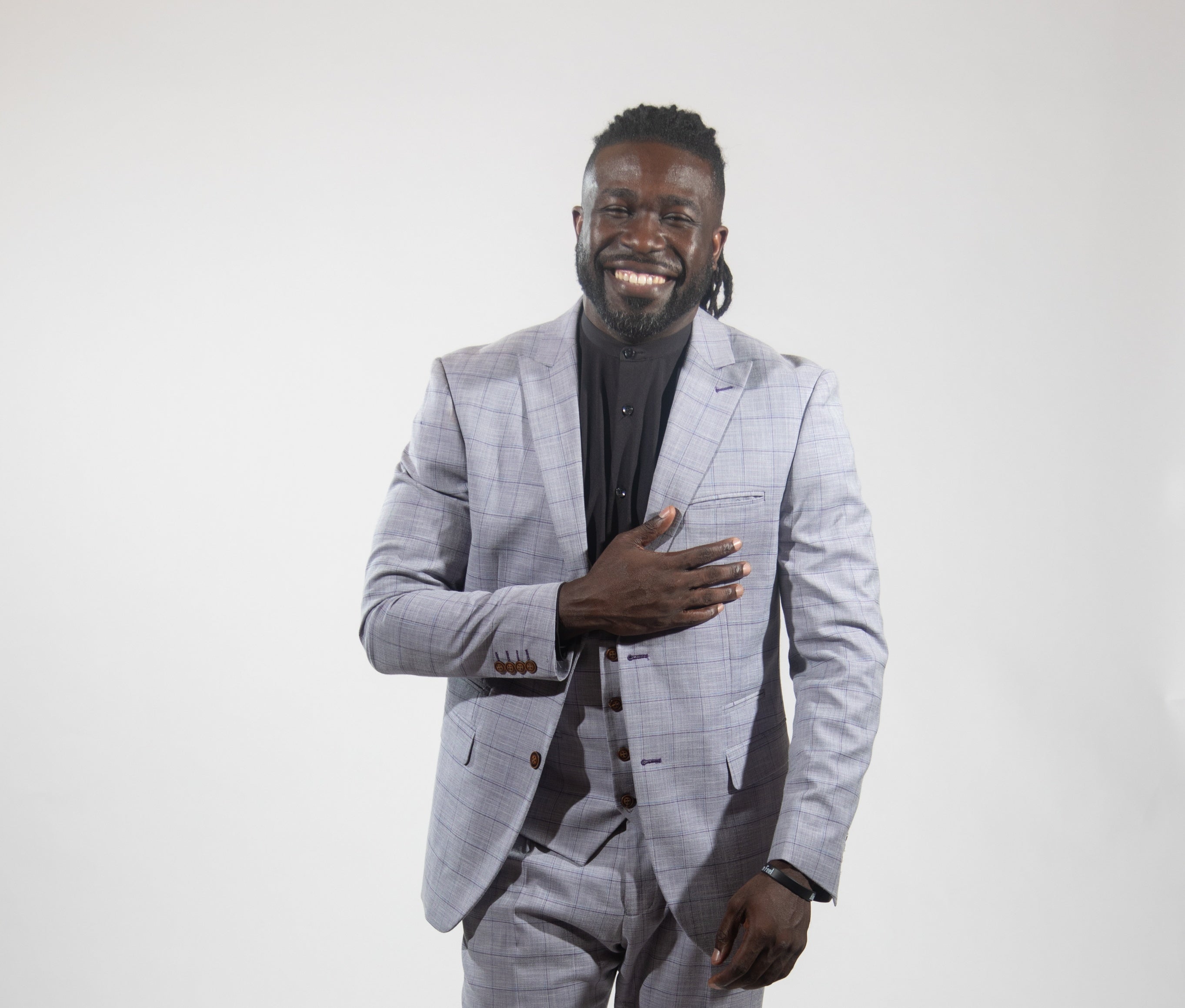 A man wears a grey suit, vest, and pants, and a black button down with one hand on his chest.