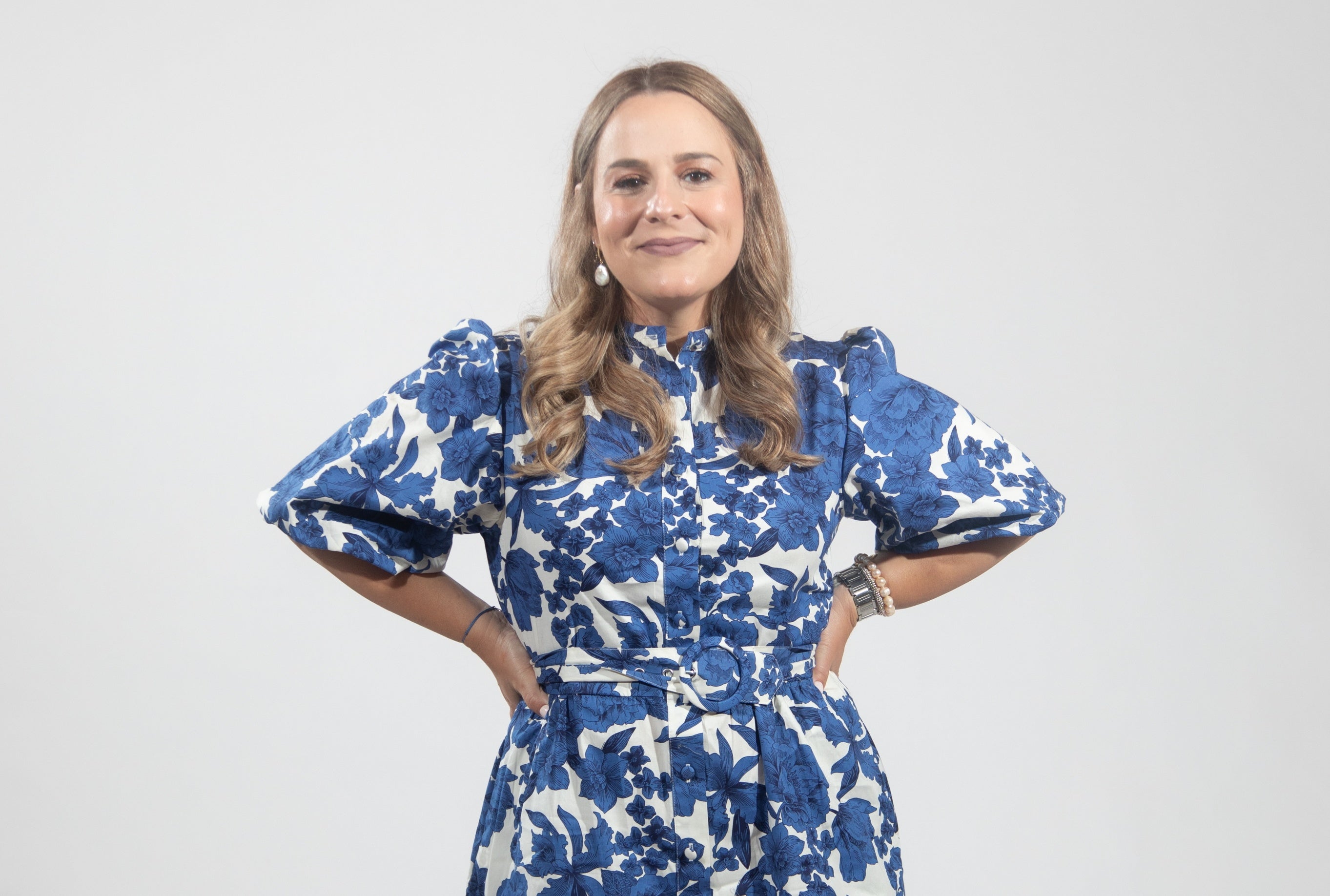 A woman with curled blonde hair wears a blue and white floral dress with elbow length sleeves.