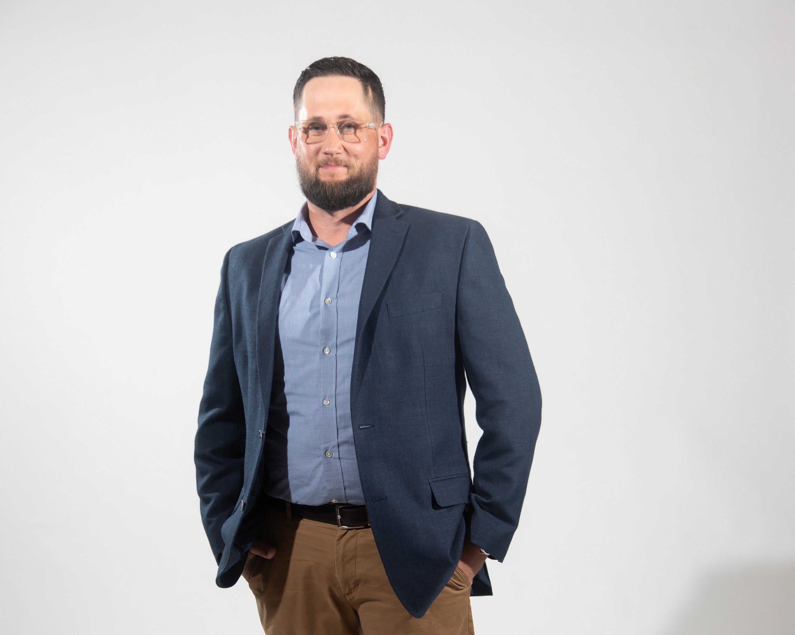 A man wears a dark blue suit jacket, light blue button down, and light brown pants with his hands in his pockets.