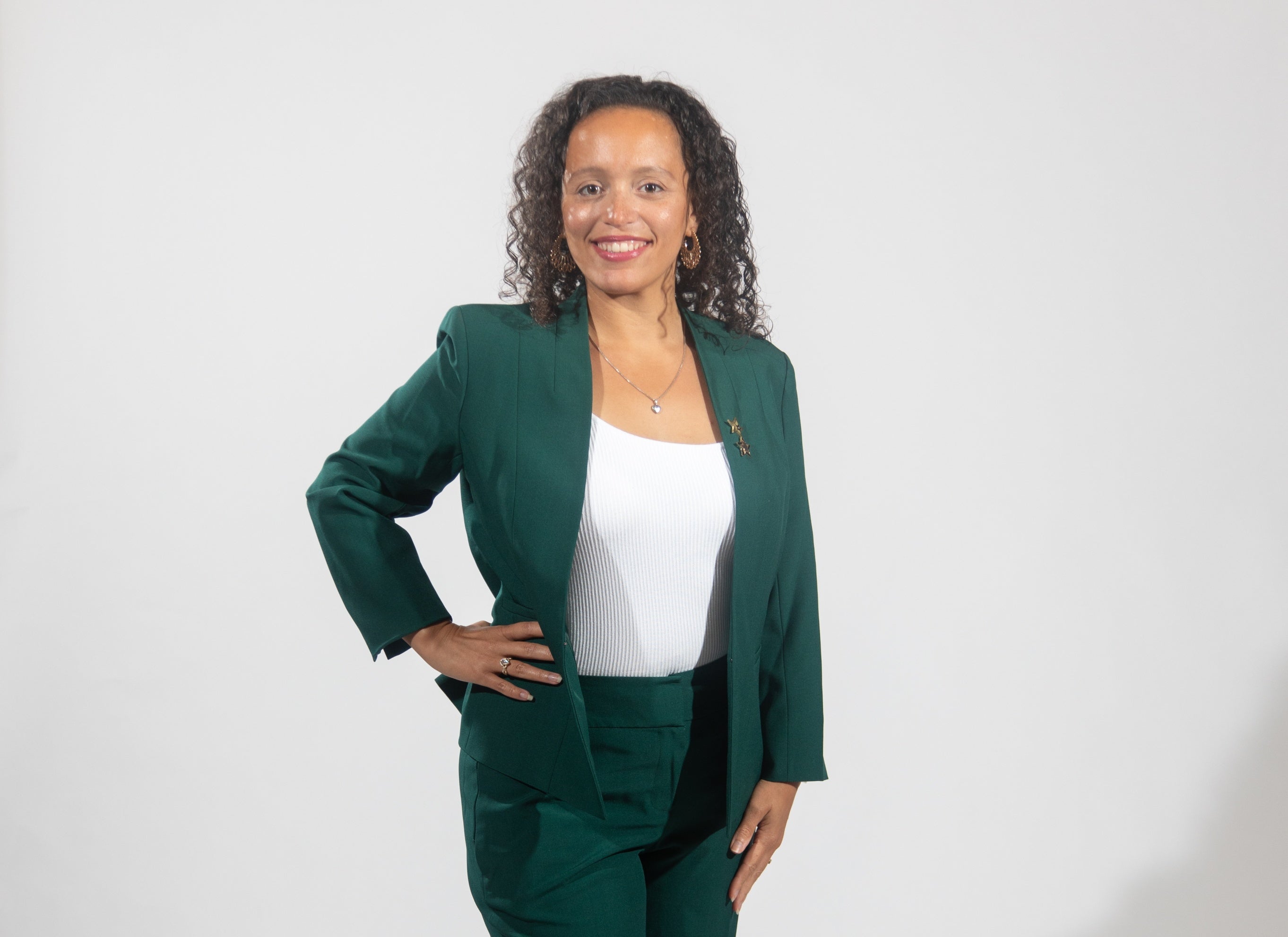 A woman with long curly hair wears a dark green suit jacket and pants and a white top.