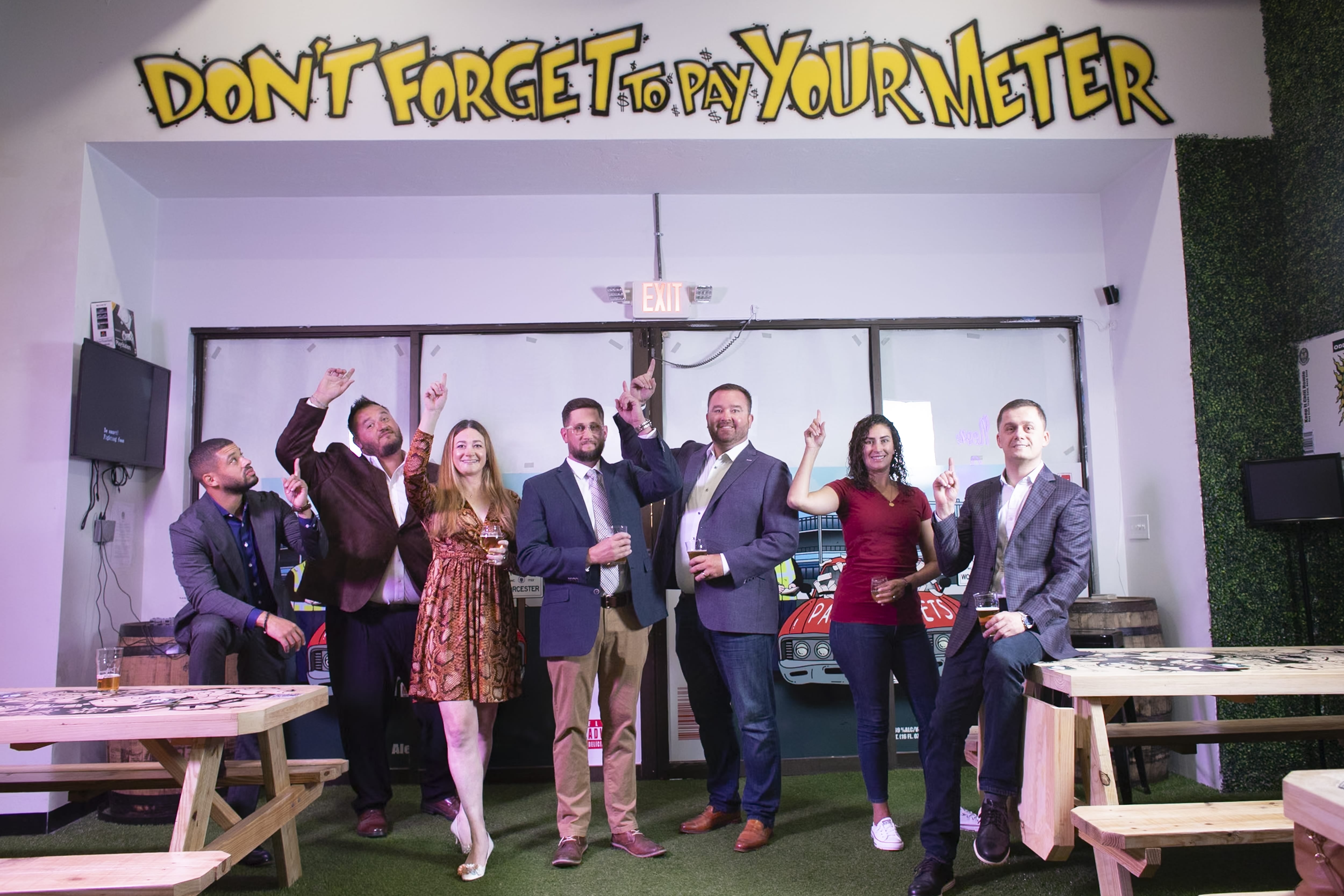 Two women and five men holding beers point at a sign above them saying "Don't forget to pay your meter!"