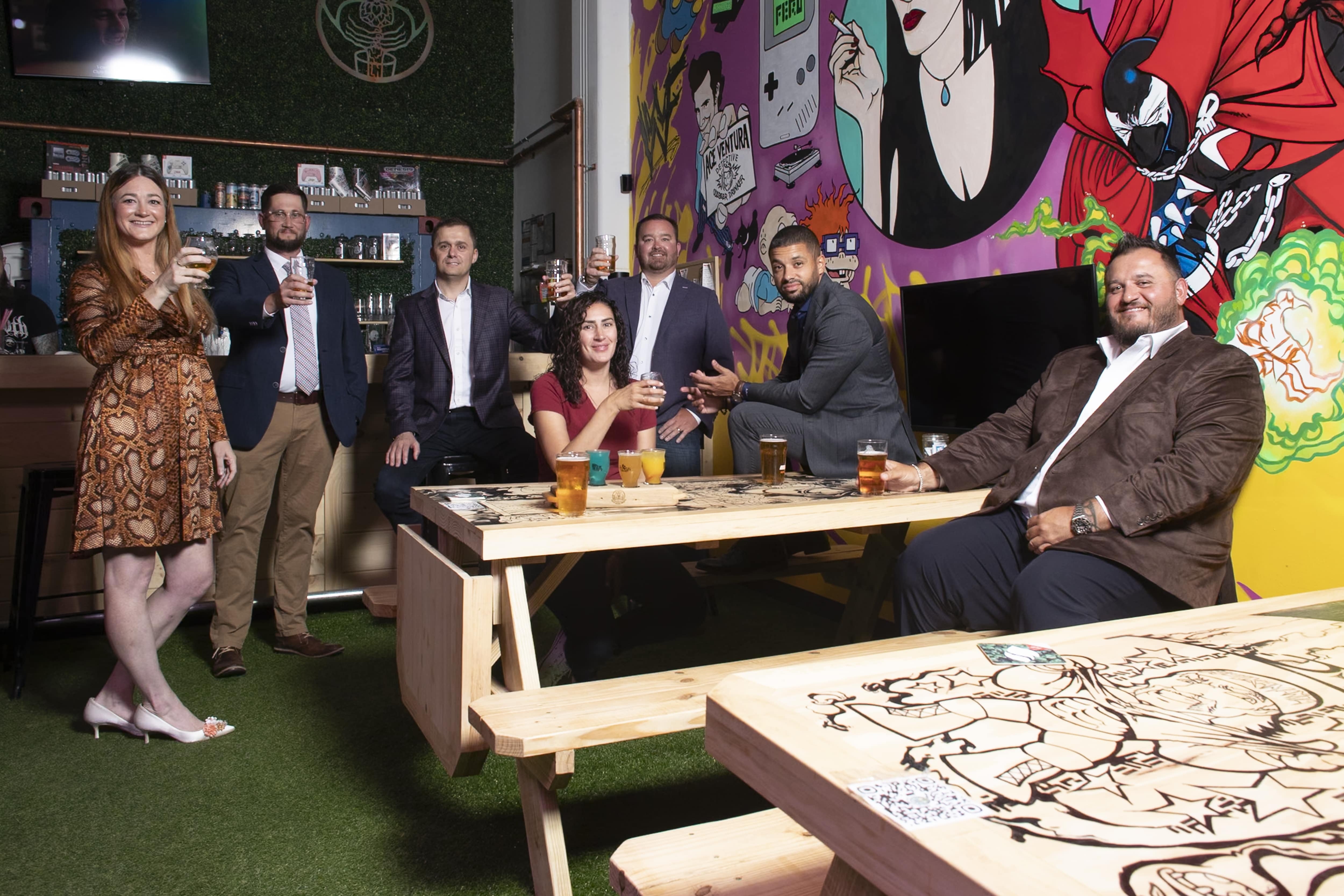 Two women and five men sit and stand in a brewery with a 1990s-themed mural in the background