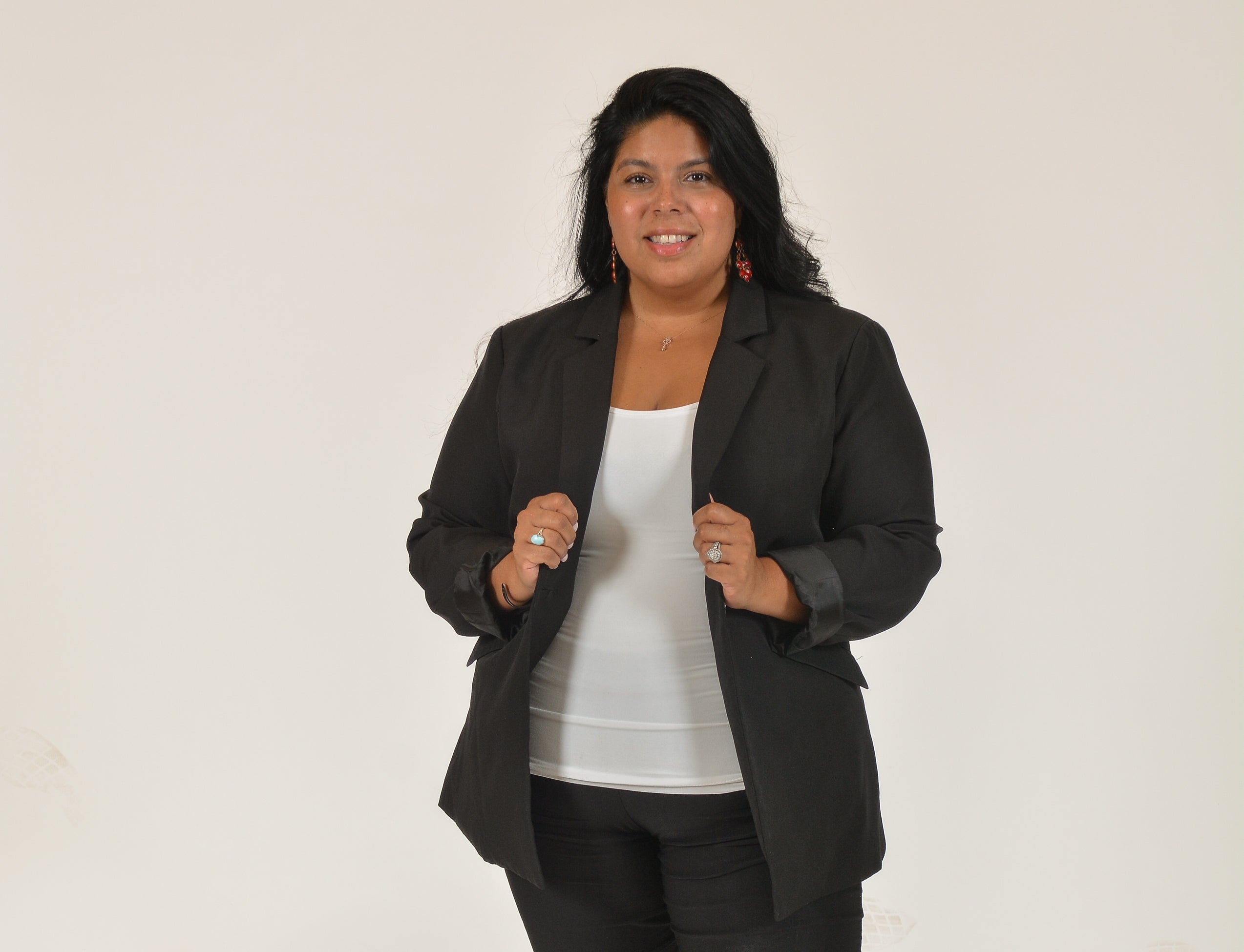 A woman with long dark hair wears a black suit jacket and pants and a white top.