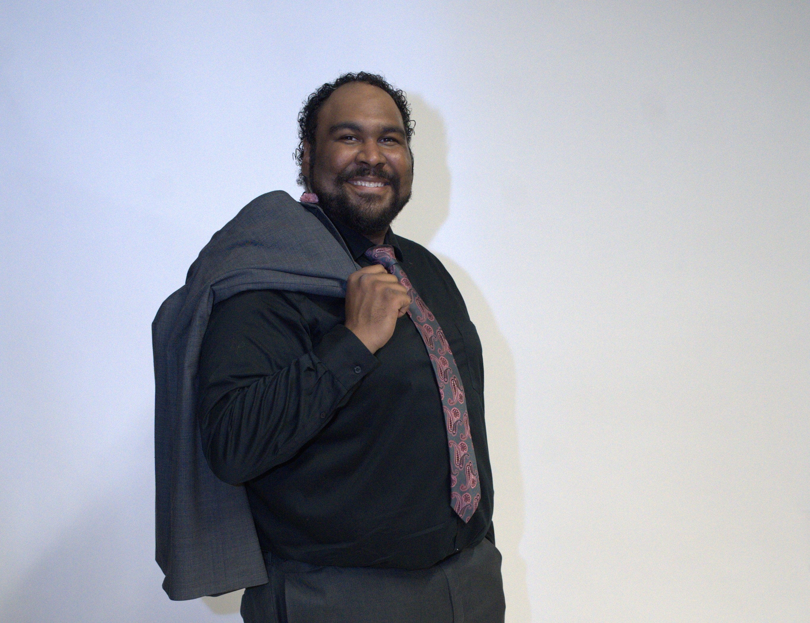 A man wears a black button down shirt, grey pants, and a grey and red paisley tie holding a grey suit jacket over his shoulder.