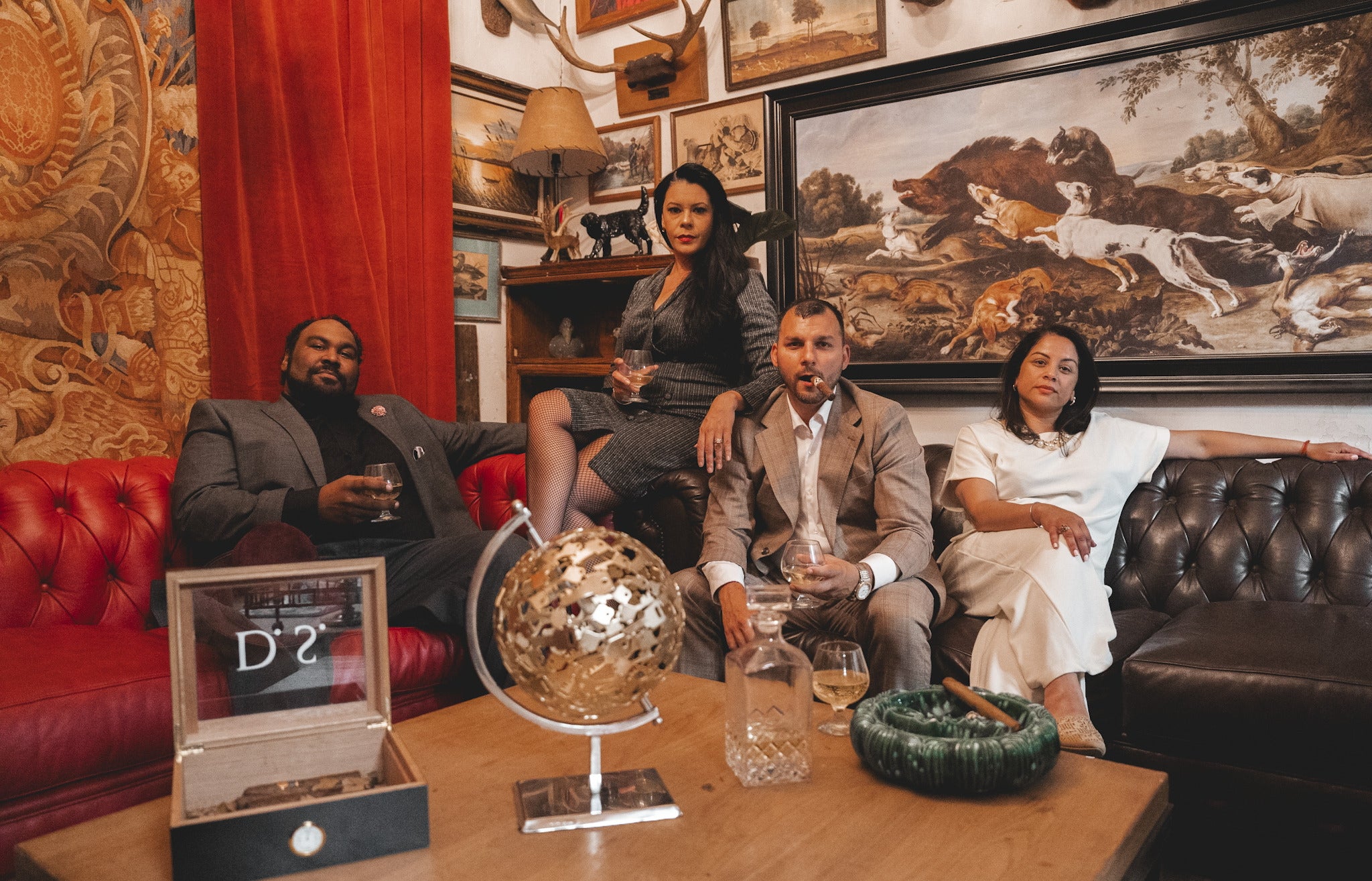 Two men and two women sit on plush couches and enjoy drinks and cigars, all of which are movie props.