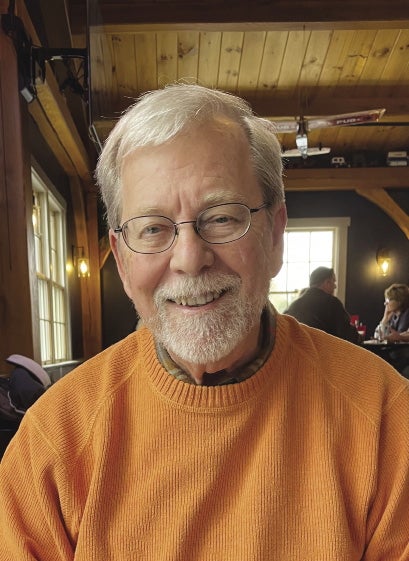 A man with grey hair and goatee beard wears an orange sweater and glasses inside a restaurant with wood panelling