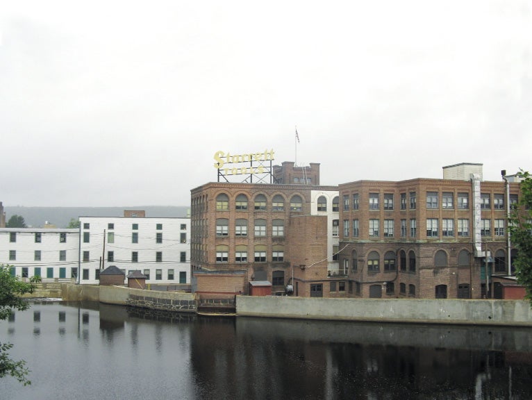 A large brick building with a yellow letters above reading "Starrett tools" sits behind a body of water in front of an overcast sky.