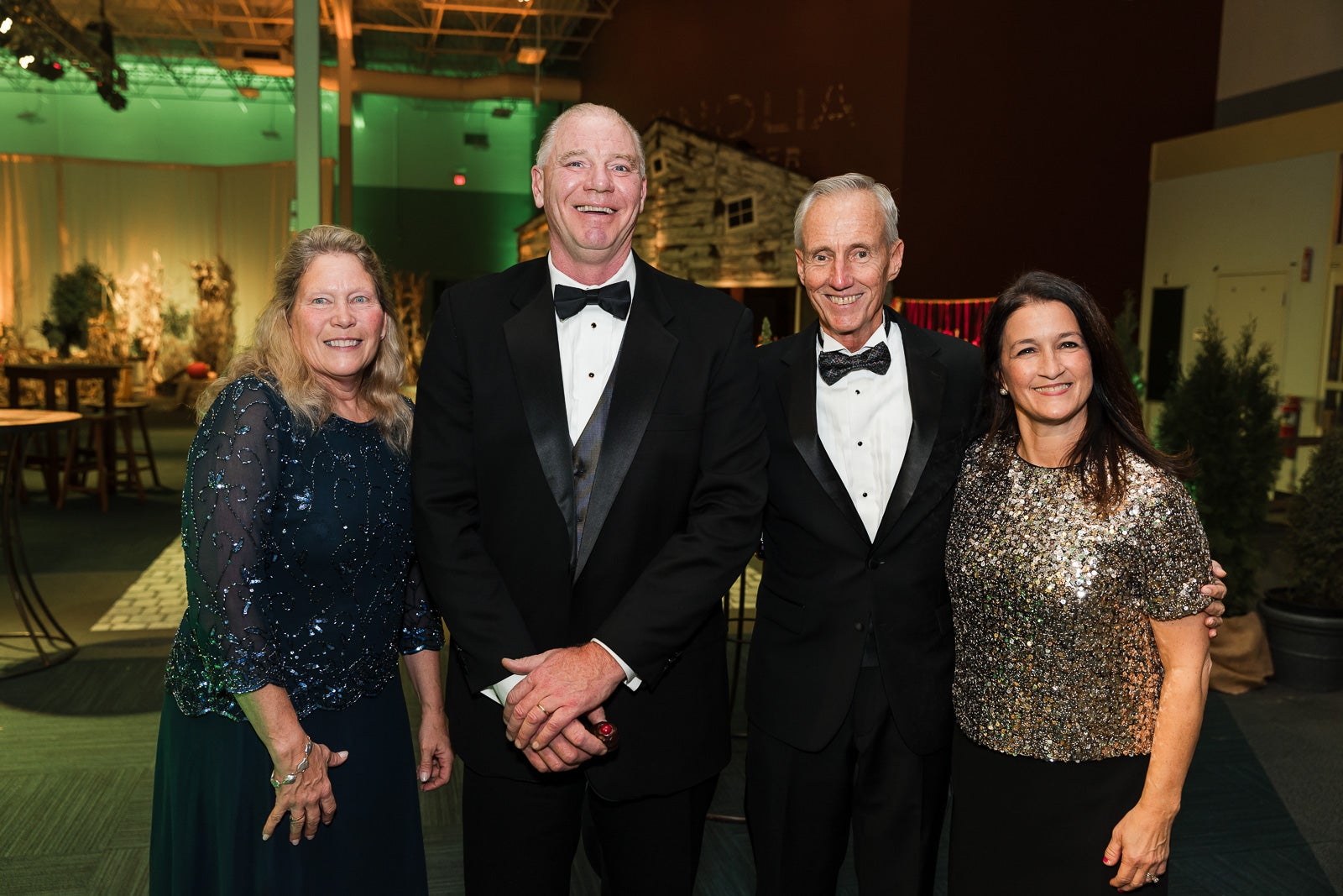 Four people stand side-by-side in an event space. The woman on the right has shoulder length blonde hair and wears a blue sparkly top and a dark blue skirt, the man to the right of her wears a black suit, the man to the right of him also wears a black suit, and the woman to the right of him wears a gold sparkly top