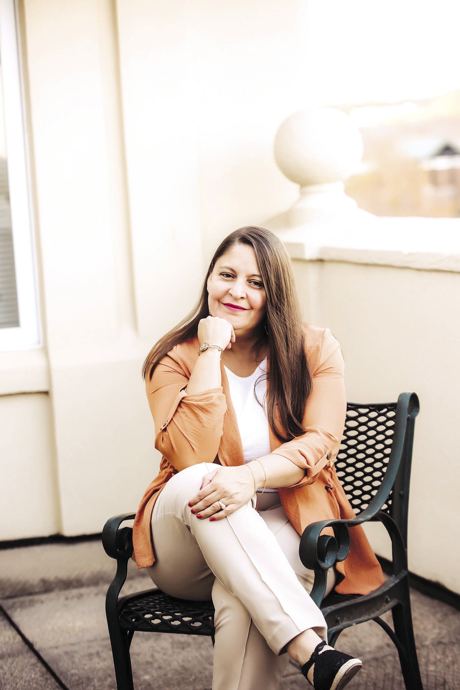 A woman with long brown hair wears a light orange cardigan, a white top, and light tan pants while sitting on a black chair with her legs crossed and her chin on her hand that rests on her thigh.