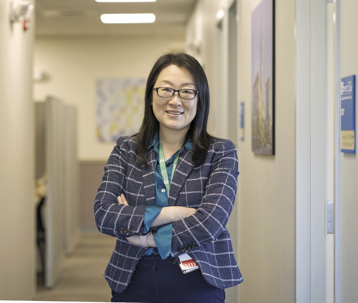 A woman with black hair wears black glasses, a blue and white patterned jacket, a teal silk top, and dark blue pants with her arms crossed standing in a hallway.