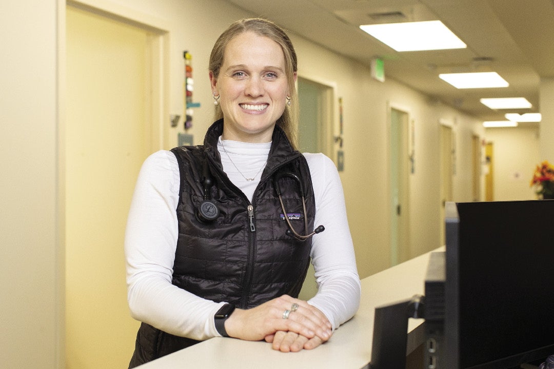 A woman with blonde hair tied back wears a long-sleeve white top, a black vest, and a stethoscope around her neck with her hands on a counter standing in a medical office hallway. 