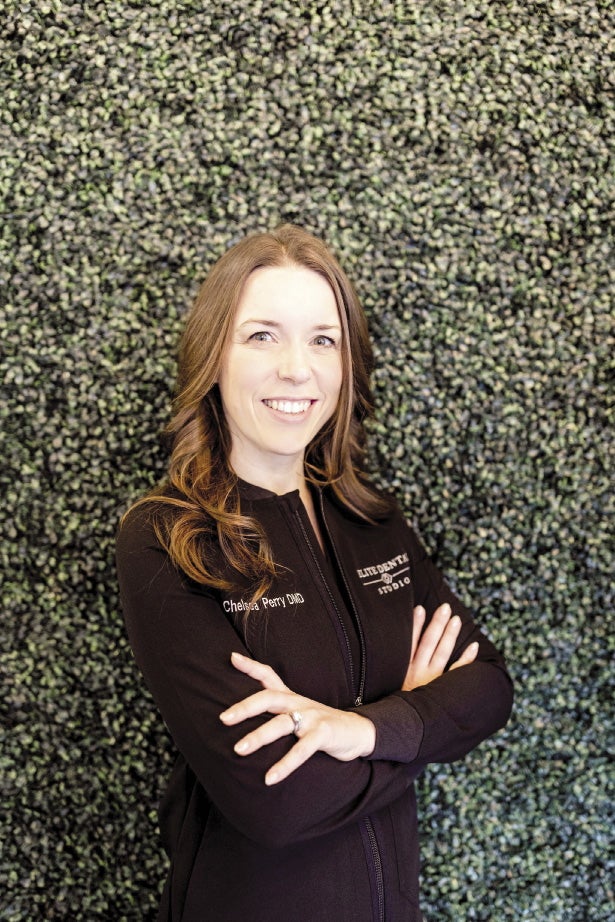 A woman with long light brown hair stands with her arms cross standing in front of a green background while wearing a black zip up jacket.