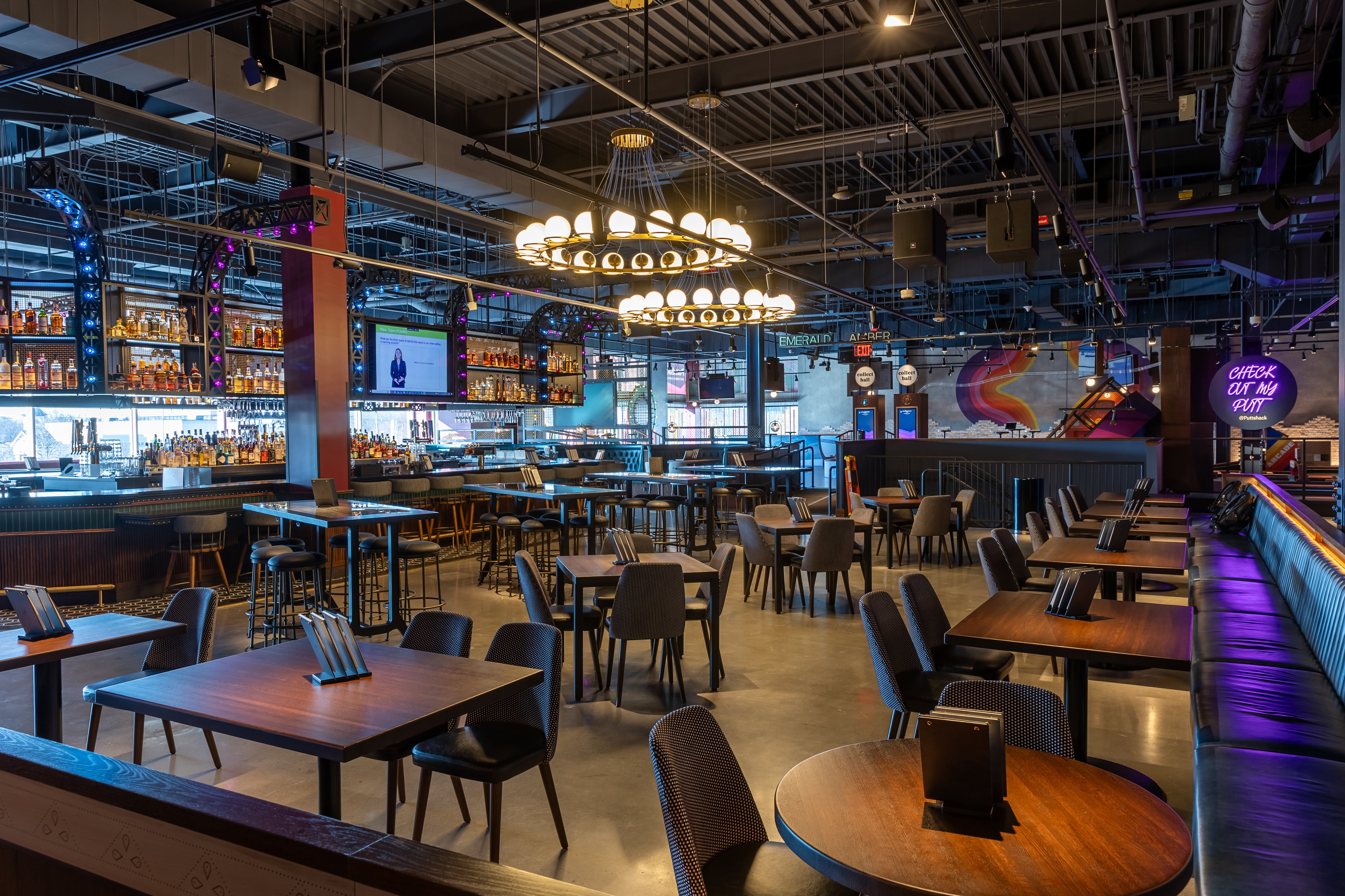 Inside a restaurant with a bar to the left, circular hanging light fixtures, and brown square and circle tables with chairs