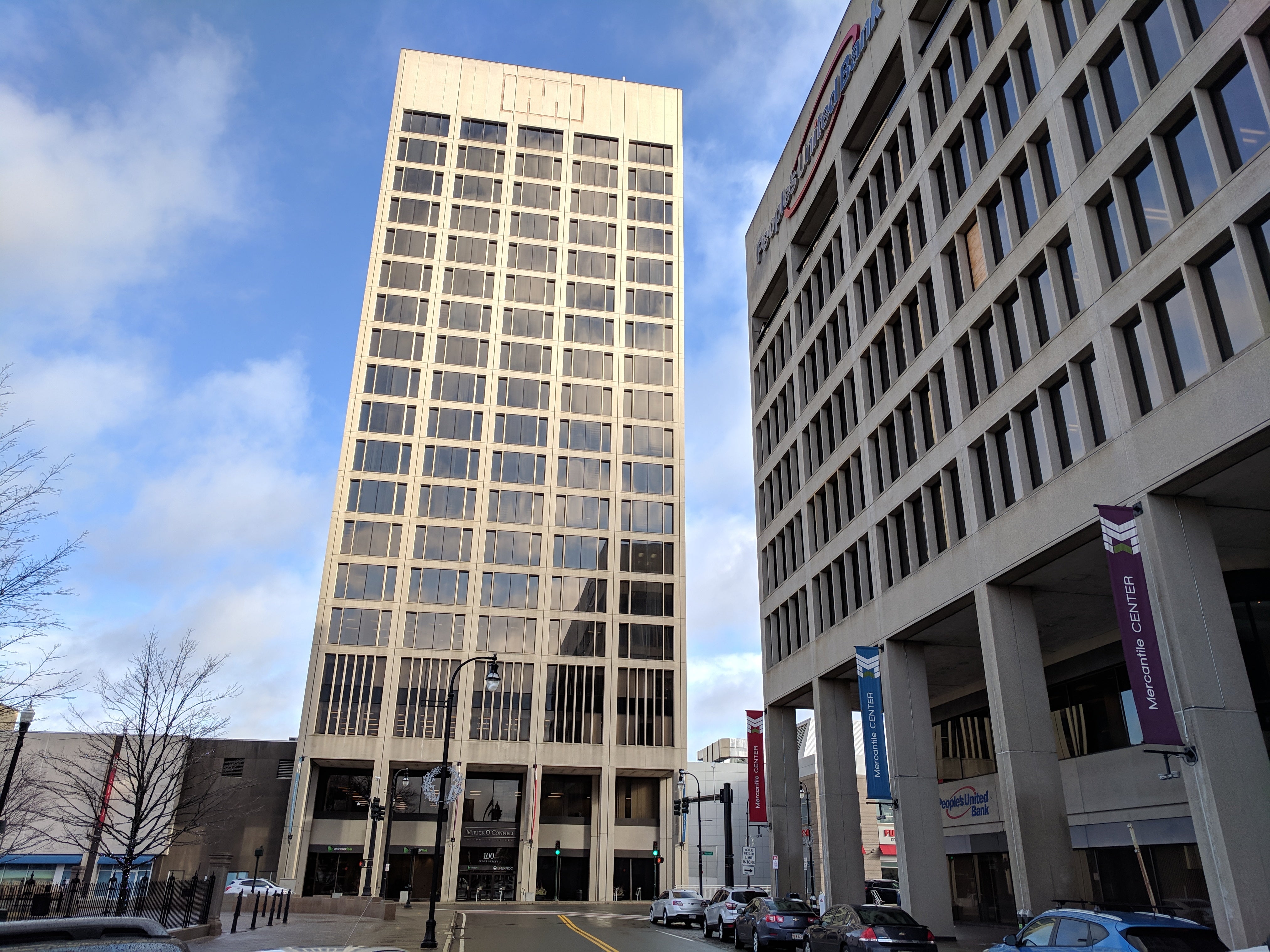 A tall building with many glass windows sits at a cross street with a blue sky with clouds behind it.