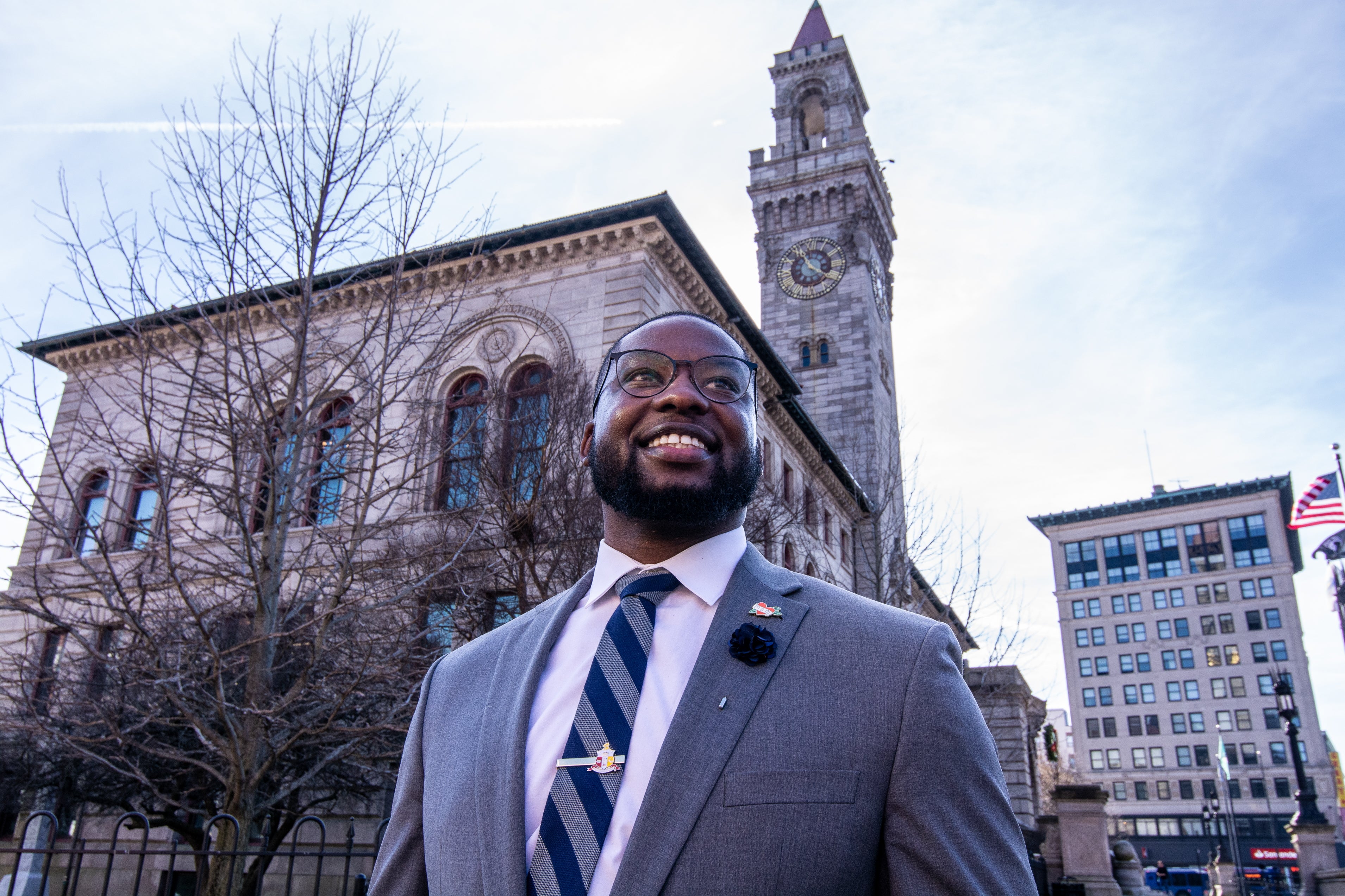 A man wears black glasses, a grey suit jacket, white button down, and stripped tie stands in front of Worcester City Hall.