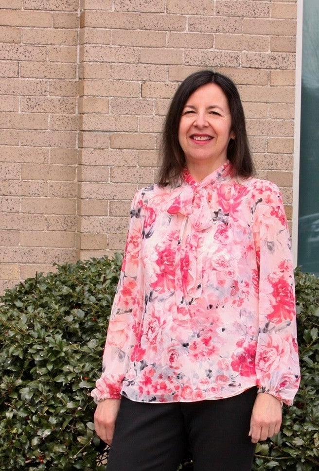 A woman with shoulder length brown hair wears a pink and white floral blouse while standing in front of shrubbery in front of a brick building.