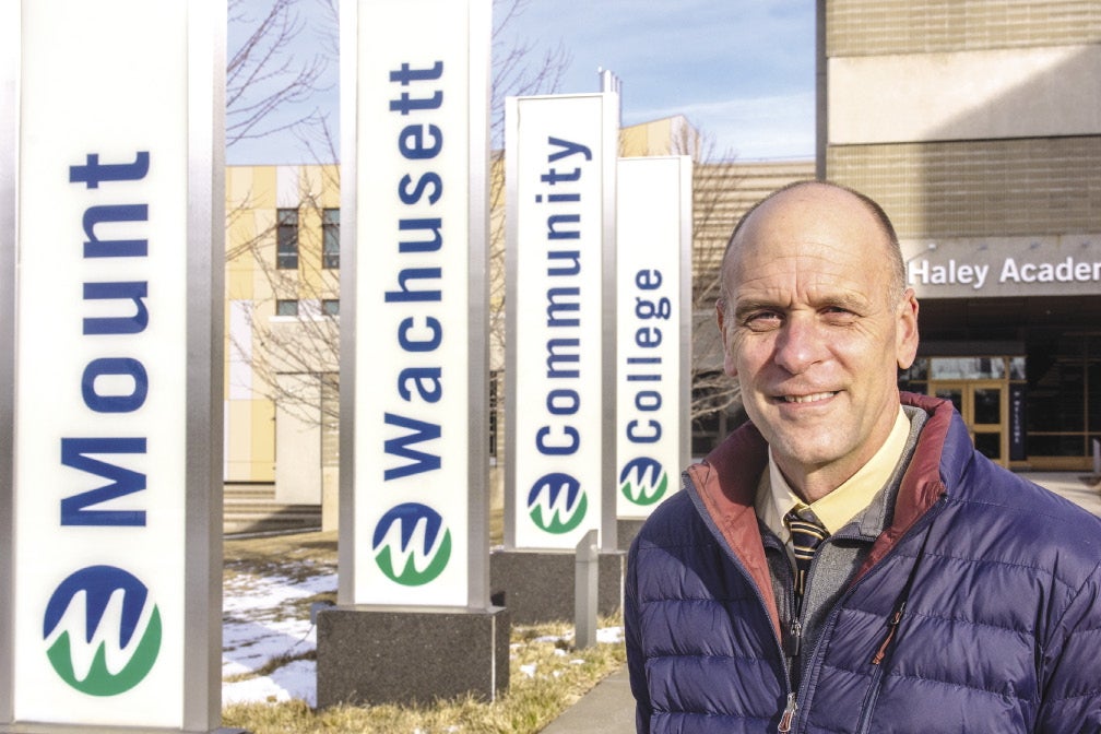 A man wears a blue puffer jacket mostly zipped up with a grey sweater, yellow button down, and striped tie while standing outside a college with four signs reading "Mount" "Wachusett" "Community" "College".