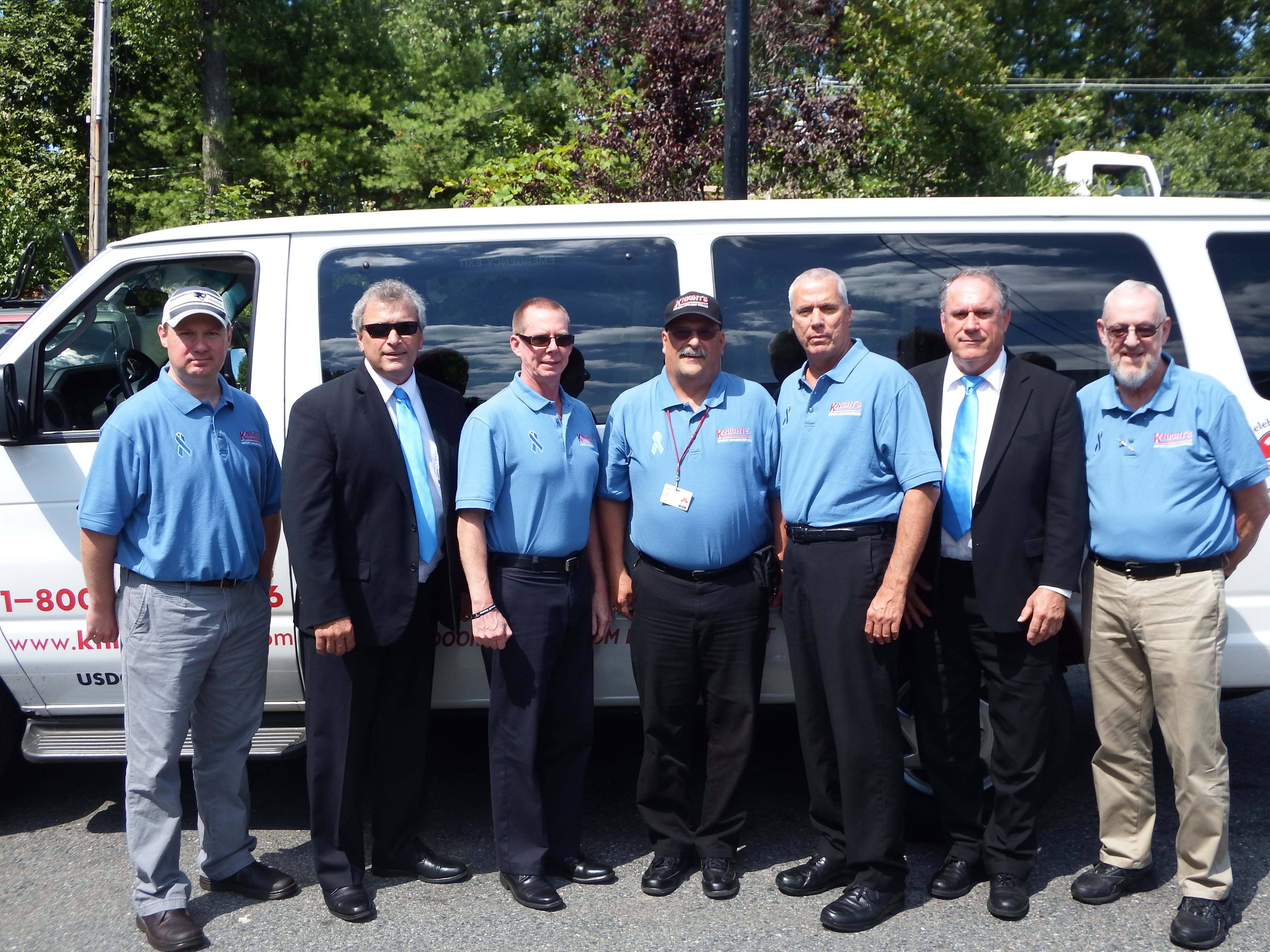 Seven men stand in line in front of a white van outside. The two on both ends of the line as well as the three in the middle wear light blue polos and the other two wear black suits, white button downs, and light blue ties.