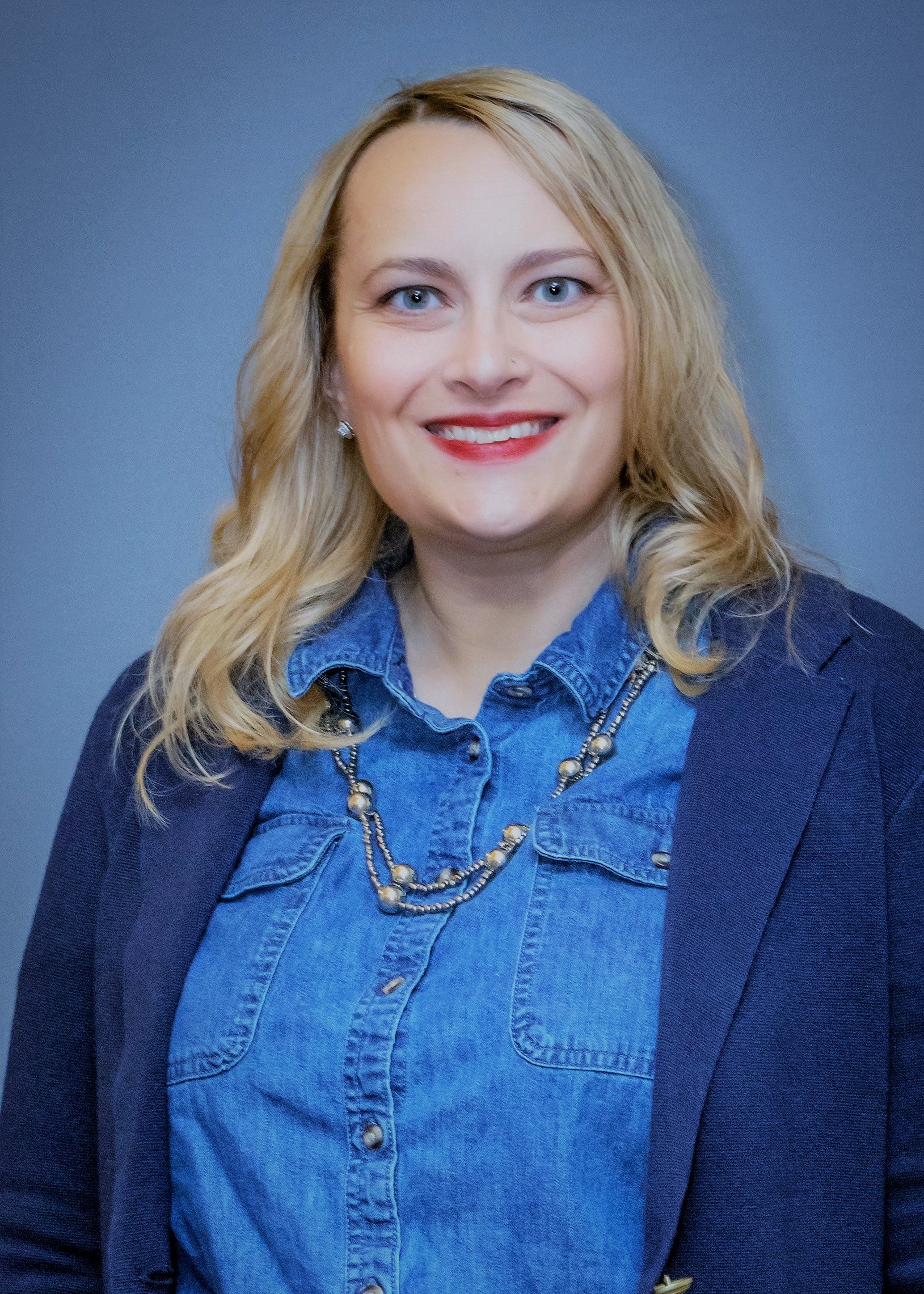 A woman with slightly curled blonde hair wears red lipstick, a blue denim top, a dark blue blazer, and a silver necklace.